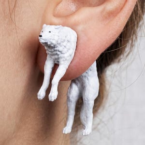 May include: White wolf earrings. These unique earrings feature a three-dimensional wolf design. The wolf's head and front legs appear to be coming out of the ear, while the body and hind legs dangle below. The earrings are a solid white color.
