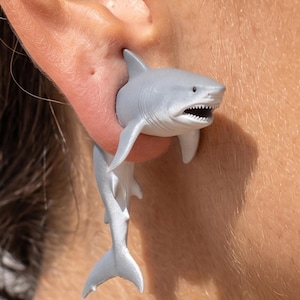 May include: A pair of gray shark earrings. The earrings are designed to appear as if a shark is biting through the ear. The shark's body extends down, with the tail visible below the ear. The earrings are a unique and playful accessory.