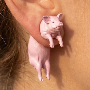 Può includere: Un paio di orecchini a forma di maiale rosa. Ogni orecchino presenta una piccola figura tridimensionale di maiale. Il maiale è in piedi, con le zampe anteriori estese. Gli orecchini sono di colore rosa chiaro e si indossano all'orecchio.