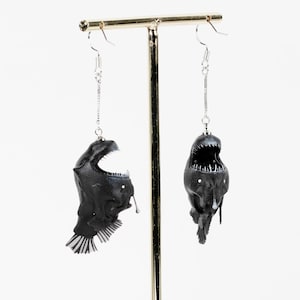 May include: A pair of black anglerfish earrings hanging from a gold stand. The earrings are made of a detailed, realistic design.