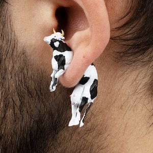 May include: A pair of novelty earrings shaped like black and white cows. The earrings have small yellow horns and pink udders. The cow earrings are worn in the ear, creating a whimsical and playful look.