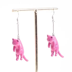 May include: A pair of dangling earrings featuring small, glossy pink cat figurines suspended from silver-toned metal chains. Each cat is depicted in a downward-facing, hanging position with its tail extended, attached to standard earring hooks.