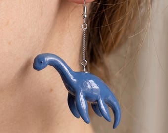 May include: A single blue dinosaur earring with a silver chain and hook. The dinosaur has a long neck and four legs. The earring is hanging from an ear.