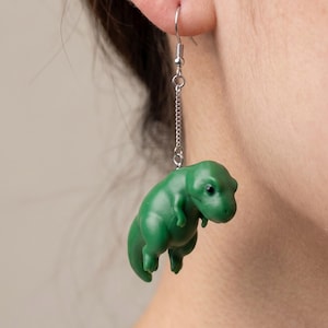 May include: A single green dinosaur earring. The earring features a small, three-dimensional Tyrannosaurus Rex charm hanging from a silver-colored chain and hook. The dinosaur is a vibrant green color with black eyes.