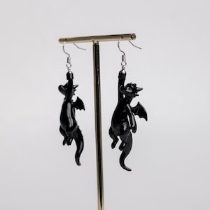 以下が含まれることがあります： 光沢のある黒いドラゴンの小さなフィギュアが付いたぶら下がりタイプのイヤリング。各ドラゴンには翼、長い尻尾、湾曲した体があり、銀色の金属製フィッシュフックワイヤーから吊り下げられているため、フィギュアは垂直にぶら下がります。