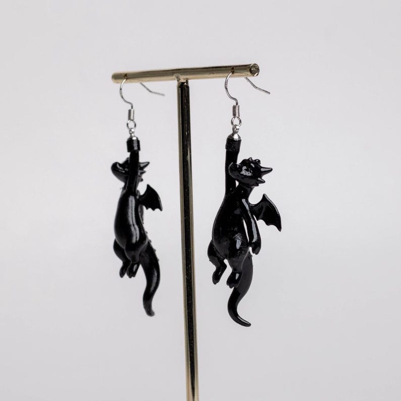 以下が含まれることがあります： 光沢のある黒いドラゴンの小さなフィギュアが付いたぶら下がりタイプのイヤリング。各ドラゴンには翼、長い尻尾、湾曲した体があり、銀色の金属製フィッシュフックワイヤーから吊り下げられているため、フィギュアは垂直にぶら下がります。