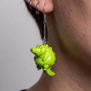 Pode incluir: Um brinco de jacaré verde-limão com corrente e gancho prateados. O jacaré tem um corpo arredondado e uma cauda pequena. O brinco é usado na orelha.