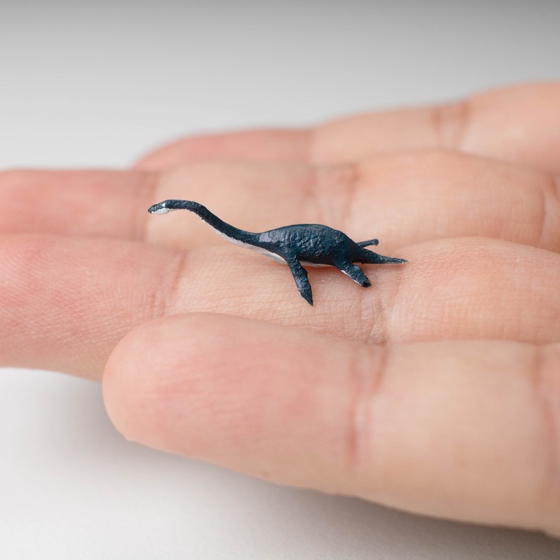 May include: A small, black, plastic dinosaur figurine of a plesiosaur, a long-necked marine reptile, on a person's hand.