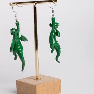 以下が含まれることがあります： 光沢のある緑色の小さなドラゴンのフィギュアが付いたぶら下がりタイプのイヤリング。各ドラゴンはぶら下がったポーズで、片腕を上に伸ばして銀色の金属製フックに固定されています。ドラゴンには翼と尻尾があります。