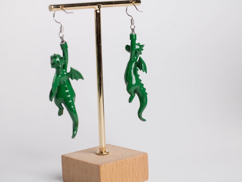 以下が含まれることがあります： 光沢のある緑色の小さなドラゴンのフィギュアが付いたぶら下がりタイプのイヤリング。各ドラゴンはぶら下がったポーズで、片腕を上に伸ばして銀色の金属製フックに固定されています。ドラゴンには翼と尻尾があります。