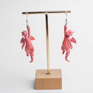以下が含まれることがあります： 小さなピンク色のドラゴンのフィギュアが付いたぶら下がりタイプのイヤリング。各ドラゴンには翼と長い尻尾があり、滑らかな質感で仕上げられています。銀色の金属製フックから吊り下げられており、片腕でぶら下がっているようなデザインです。