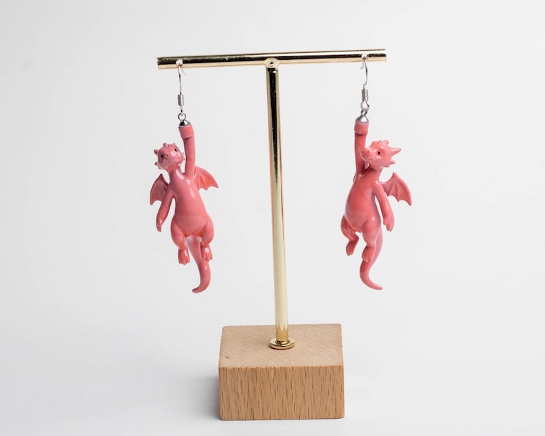 以下が含まれることがあります： 小さなピンク色のドラゴンのフィギュアが付いたぶら下がりタイプのイヤリング。各ドラゴンには翼と長い尻尾があり、滑らかな質感で仕上げられています。銀色の金属製フックから吊り下げられており、片腕でぶら下がっているようなデザインです。