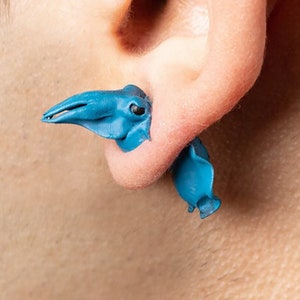 Könnte beinhalten: Ein Paar blaue Tintenfisch-Ohrringe. Die Ohrringe sind aus einem weichen, biegsamen Material gefertigt und haben die Form eines Tintenfischs mit einem langen, dünnen Schnabel.