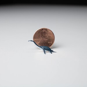 May include: A small, black and white plastic dinosaur figurine next to a US penny. The penny is dated 2016 and has the words "In God We Trust" and "Liberty" on it.