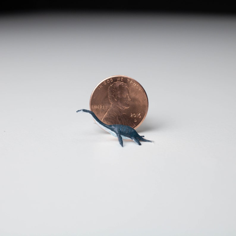 May include: A small, black and white plastic dinosaur figurine next to a US penny. The penny is dated 2016 and has the words "In God We Trust" and "Liberty" on it.
