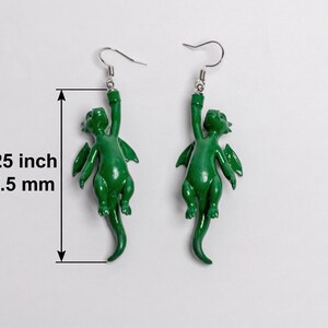 以下が含まれることがあります： 銀色のフックが付いた緑色のドラゴン型ダングルピアス一対。各ピアスには、片腕でぶら下がっている小さな緑色のドラゴンのフィギュアがデザインされています。測定表示により、ドラゴンのフィギュアの全長が57.5mmであることが示されています。