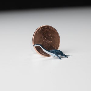 May include: A small, blue and white dinosaur figurine next to a US penny. The penny is dated 2016 and has the words "In God We Trust" and "Liberty" on it.