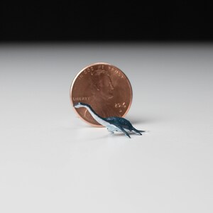 May include: A small, blue and white dinosaur figurine next to a US penny. The penny is dated 2016 and has the words "In God We Trust" and "Liberty" on it.