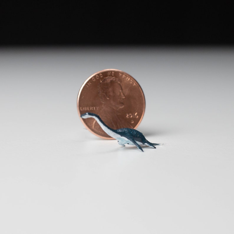 May include: A small, blue and white dinosaur figurine next to a US penny. The penny is dated 2016 and has the words "In God We Trust" and "Liberty" on it.