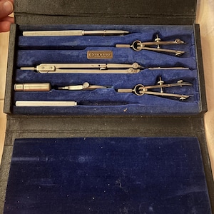 以下が含まれることがあります： 青いベルベットの内装と、いくつかのヴィンテージ製図ツールが入った、開いた黒い革ケース。ツールには、コンパス、ディバイダー、鉛筆ホルダーが含まれます。ケースは開いており、ツールがきちんと配置されています。