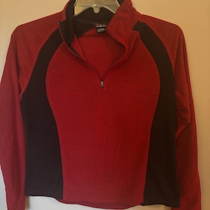 May include: A red and black long-sleeved athletic top with a half-zip closure. The top features a textured, waffle-like pattern in red, with black panels on the sides. The collar and zipper are black.