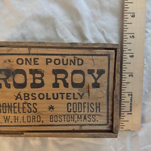 May include: A rectangular wooden box with black printed text reading ONE POUND ROB ROY ABSOLUTELY BONELESS CODFISH W.H.LORD, BOSTON, MASS. A wooden ruler is placed vertically next to the box, showing a height of approximately 15 centimetres.