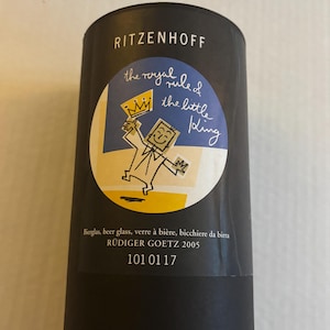 May include: A black cylindrical container with a circular label featuring a cartoon king holding a crown. The text "RITZENHOFF" is at the top. The label reads "the royal rule of the little king". Below the label are the words "Bierglas, beer glass, verre à bière, bicchiere da birra".