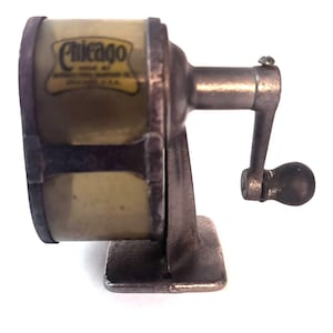 Puede incluir: Un sacapuntas manual vintage con acabado metálico oscuro y cuerpo cilíndrico. El lateral presenta una etiqueta con el texto Chicago. Incluye una manivela con un pomo negro redondo y una base cuadrada para fijar sobre una superficie plana.