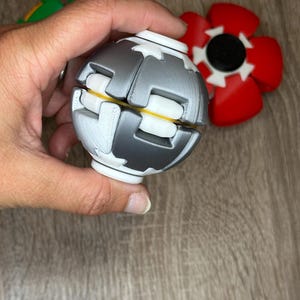 Könnte beinhalten: Ein kugelförmiges Fidget-Spielzeug mit ineinandergreifenden grauen und weißen Kunststoffsegmenten und einem gelben Innenkern. Das Spielzeug hat ein glattes, abgerundetes Design mit sichtbaren 3D-Druckschichten. Weitere rote und grüne Spielzeuge sind sichtbar.