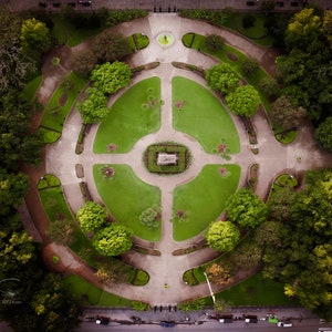 Könnte beinhalten: Eine Luftaufnahme eines kreisförmigen Parks mit einem zentralen Springbrunnen und konzentrischen Wegen, die von Bäumen gesäumt sind. Der Park ist von Gebäuden und einer Straße umgeben.