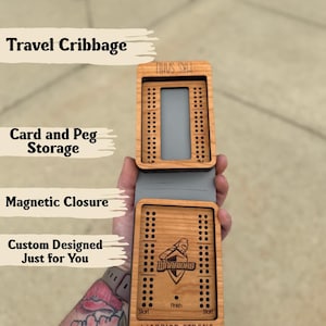 May include: A wooden travel cribbage board with a magnetic closure. The board has card and peg storage and is custom designed. The text "WARRIOR STRONG" and "WARRIORS" are visible on the board.