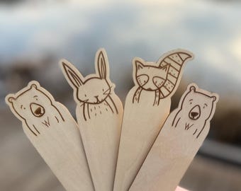 May include: Four wooden bookmarks with different animal designs. The bookmarks are light brown and feature a bear, a rabbit, a raccoon, and another bear. Each bookmark has a small, decorative design at the bottom.