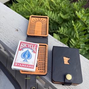 Könnte beinhalten: Ein tragbares Cribbage-Spielbrett aus Holz mit einem schwarzen lederartigen Einband, der einen Ausschnitt des Bundesstaates Minnesota aufweist. Das Set enthält ein Kartenspiel von Bicycle Standard und einen Messing-Druckknopfverschluss.