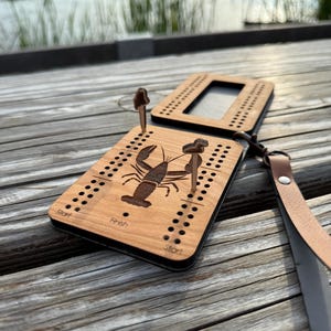 Könnte beinhalten: Ein hölzernes Cribbage-Spielbrett mit einem lasergravierten Hummer-Design. Das Brett hat ein Scharnierdesign, Stifte und einen Lederriemen. Die Wörter "Start" und "Finish" sind auf dem Brett sichtbar.