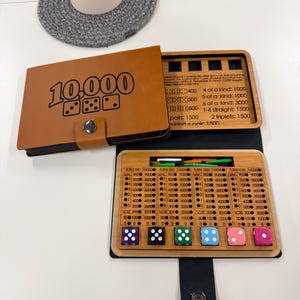 Könnte beinhalten: Ein braunes, lederbezogenes Würfelspiel mit dem Titel 10.000 und einem hölzernen Punktezähler für vier Spieler. Das Set enthält sechs bunte Würfel, kleine Spielsteine aus Kunststoff und eine aufgedruckte Spielanleitung auf dem Innendeckel.