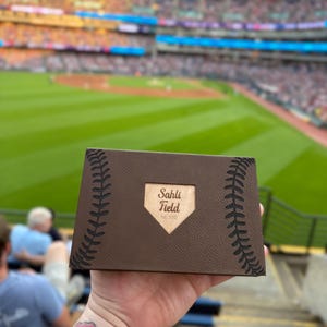 May include: A brown leather box with baseball stitching and a wooden home plate design that reads "Sahli Field Est. 2012". The box is held in front of a baseball stadium.