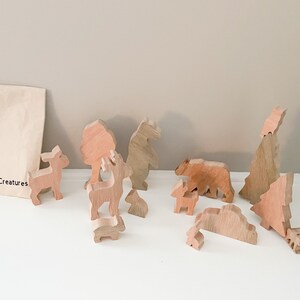 May include: Wooden woodland creature toys, including a deer, bear, and trees, are arranged on a white surface. A canvas bag with the text "Woodland Creatures" is visible. The toys are made of light-colored wood.
