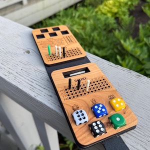 Puede incluir: Un tablero de juego de dados de viaje de madera con dos secciones de puntuación con pequeñas clavijas, la palabra SCORE y seis dados coloridos en blanco, azul, amarillo, verde y negro. El tablero incluye iconos grabados y una correa de cuero negro.