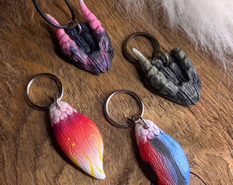 Puede incluir: Cuatro accesorios esculpidos que incluyen un colgante con cuernos negro y rosa en un cordón, un llavero de garra gris oscuro texturizado, un llavero con forma de hoja degradado rojo y amarillo, y otro rojo, negro y azul.