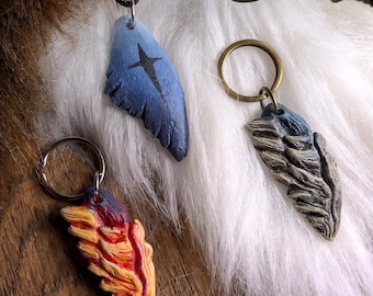 Puede incluir: Tres accesorios esculpidos en forma de ala: un colgante azul con un símbolo de estrella negra en un cordón negro, un llavero gris y azul, y un llavero rojo, naranja y amarillo. Cada pieza presenta detalles de plumas texturizados.