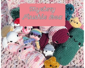 Puede incluir: Una colección de pequeños peluches de ganchillo de diversos colores y formas, incluyendo un conejo rosa, un pepinillo verde, un pulpo arcoíris y estrellas amarillas, con un cartel rosa que dice Mystery Plushie Box.