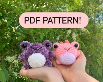 May include: Two small crocheted frog plushies, one purple with a white belly and one pink with a lighter pink belly, both featuring large black plastic eyes and stitched mouths. The image includes the text PDF PATTERN in a pink rounded box.