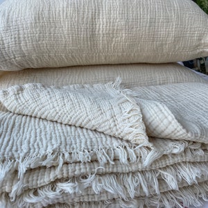 Op de afbeelding: Een stapel witte katoenen dekens met een gefranst randje. De dekens zijn gevouwen en op elkaar gelegd, wat een zachte en knusse uitstraling geeft.