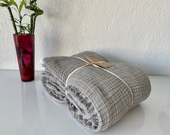 May include: A gray, woven throw blanket with fringed edges is folded and tied with a white ribbon. The blanket is on a white surface.