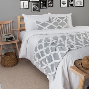 May include: A white bed with a gray and white patterned throw blanket. The bed has a white headboard and two white pillows with a gray and white patterned design. The bed is in a bedroom with a gray wall and a wooden floor. There is a wooden chair with a typewriter on it and a wicker basket on the floor.