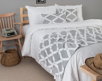 Può includere: Un letto bianco con una coperta a motivi grigi e bianchi. Il letto ha una testiera bianca e due cuscini bianchi con un motivo a griglia grigia e bianca. Il letto si trova in una camera da letto con una parete grigia e un pavimento in legno. C'è una sedia di legno con una macchina da scrivere sopra e un cesto di vimini sul pavimento.