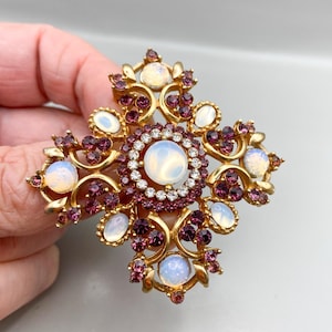 May include: A gold-toned cross-shaped brooch with a central large, round, opalescent stone surrounded by a ring of clear crystals. The cross arms feature smaller opalescent stones and clusters of deep purple crystals. The brooch is held by a hand.