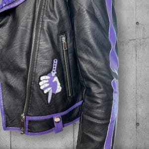 May include: Black faux leather jacket with purple accents. Features a zipper closure, a small pocket, and a decorative patch of a skeleton hand holding a purple dagger. The jacket has purple trim and a unique design on the sleeve.