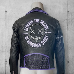 May include: Black leather jacket with purple accents and the text "ALWAYS THE VILLAIN IN SOMEONES STORY" on the back. The jacket features a graphic of a smiling mask. The collar and lower back panel have purple detailing.