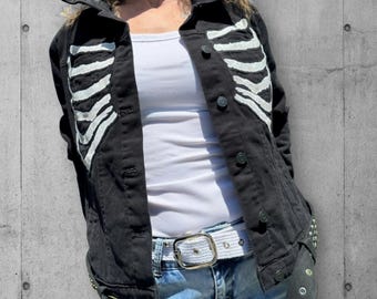 May include: A black denim jacket with a white painted skeleton ribcage design on the front. The jacket has a button closure and a white belt.